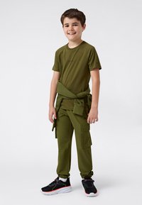 Grønn treningsantrekk bestående av en t-skjorte og cargo-bukser. T-skjorten har et logo, buksene har lommer. Joggeskoene er svarte med neonlys.