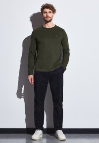 Camiseta de manga larga en color verde oliva combinada con pantalones negros y zapatillas blancas, con cuello redondo y un logo sutil en el pecho.