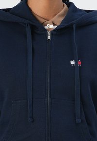 Felpa con zip navy realizzata in tessuto morbido, dotata di una tasca frontale, cordini e una piccola etichetta con logo nei colori rosso, bianco e blu.