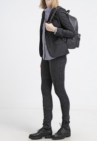 Leggings neri aderenti con dettagli texturizzati, un blazer nero sopra una camicia a quadri e stivali neri chunky con lacci. Zaino nero indossato.