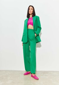 Groen op maat gemaakt pak met wijde broekspijpen en een lange blazer, gecombineerd met een roze cropped top en roze instappers met hardware detail.