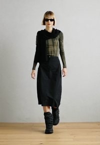 Svart skjørt med asymmetrisk design, kombinert med en grønn langermet topp med et tie-dye mønster. En svart skjerf og kraftige svarte støvler fullfører antrekket.