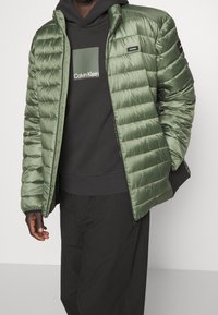 Persoon draagt een opengewerkte olijfgroene gewatteerde jas over een zwarte Calvin Klein hoodie en losse zwarte broek, met één hand in de jaszak.