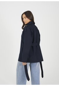 Granatowy trencz z paskiem w talii, guzikami i kołnierzykiem. Materiał wydaje się gładki, ma dopasowany krój oraz boczne kieszenie.