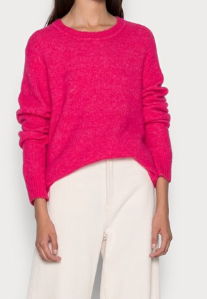Femme portant un pull douillet à manches longues rose vif et un pantalon ample blanc cassé, les mains détendues de chaque côté.