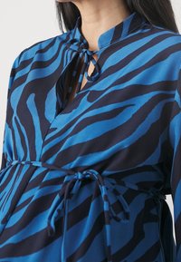 Chemise rayée bleu et noir avec un col en trou de serrure, fermeture par lien et coupe décontractée. Fabriquée dans un tissu lisse à la texture légère.