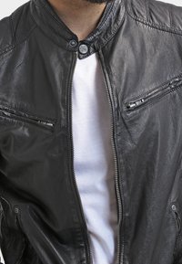 Schwarze Lederjacke mit hohem Kragen, Reißverschluss vorne und zwei Reißverschlusstaschen auf der Brust. Glatte Textur mit dezenten Nähten.