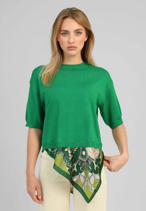 Femme blonde portant un haut vert à manches courtes sur une écharpe verte à motifs et un pantalon crème, debout devant un fond gris.