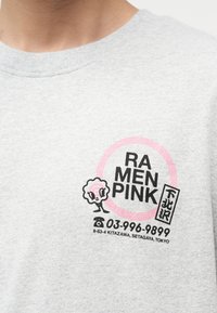 Γκρι μπλουζάκι με μαύρο και ροζ λογότυπο καταστήματος ράμεν που απεικονίζει καρτούν μπολ με ράμεν, αριθμό τηλεφώνου και διεύθυνση στο Τόκιο στο στήθος.