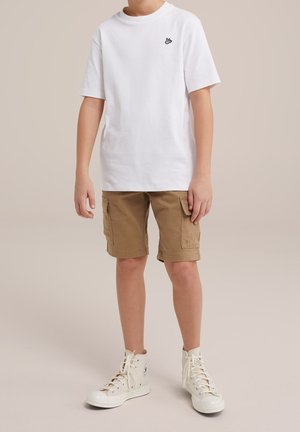 Enfant portant un t-shirt blanc à manches courtes avec un petit logo noir, un short cargo beige, et des baskets blanches montantes, debout sur un fond neutre.
