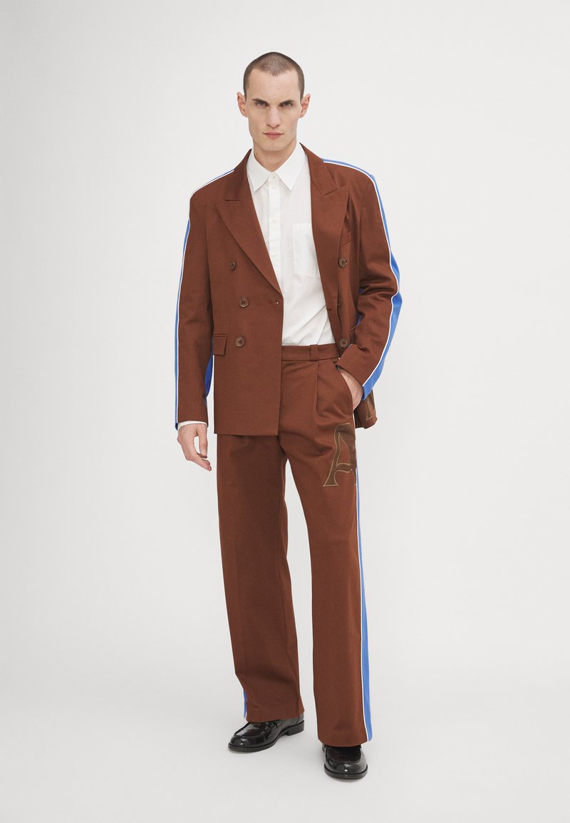 Blazer marrone doppiopetto con strisce laterali blu, abbinato a pantaloni coordinati con una grafica dorata "A". Camicia bianca sotto.