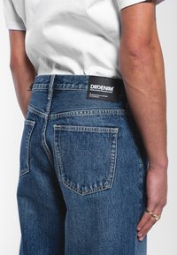 Blue denim jeans with a high waist, featuring prominent back pockets and a black label at the waistband. Texture appears durable and classic.