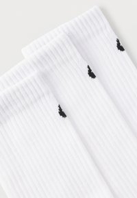 Τρία ζευγάρια λευκές κάλτσες μέσης με ριγέ υφή και μικρά μαύρα σχέδια καρδιάς κοντά στις επάνω μανσέτες, τοποθετημένες αλληλοκαλυπτόμενες σε λευκή επιφάνεια.