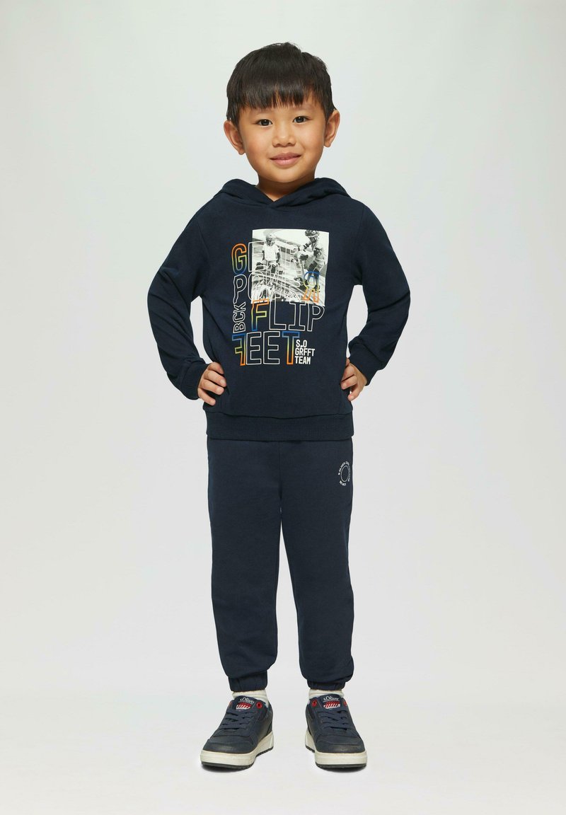 Haut à capuche bleu marine avec impression graphique et texte, assorti à un pantalon de survêtement. Comprend des poignets élastiques et des baskets décontractées.