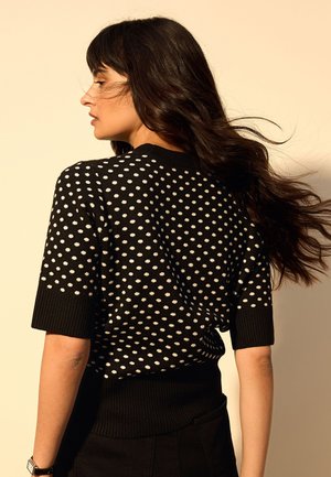 Donna con lunghi capelli scuri girata di lato, indossa un maglione nero a maniche corte con pois bianchi su sfondo beige.