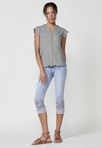 Graue bestickte Bluse mit kurzen Ärmeln und Knopfleiste; hellblaue cropped Jeans mit aufwendiger floralem Stickmuster am Saum; beigefarbene Sandalen mit Riemen.