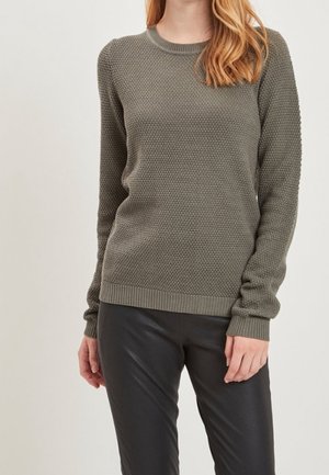 Pull en tricot vert kaki avec un motif texturé, col rond et manches longues, associé à un pantalon en cuir noir. Poignets et ourlet côtelés.