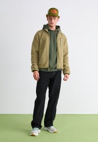 Fleece-takk i olivengrønn, lagd over en mørkere grønn hettegenser. Svart bukser, grå joggesko, og en grønn caps med en oransje logo.