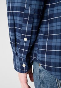 Camisa de cuadros de manga larga con tonos azules, patrón de cuadrícula blanca, puños abotonados y dobladillo curvado, hecha de una tela texturizada.