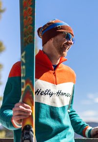 Kolorowy sweter Helly Hansen w odcieniach pomarańczowym, białym i turkusowym, z zamkiem przy kołnierzu. Do stroju pasuje jasnobrązowa dziana czapka i okulary przeciwsłoneczne.