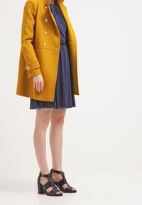 Abrigo mostaza con botones plateados sobre un vestido azul, complementado con sandalias de tacón alto negras con tiras. Tejido suave y ajuste entallado.