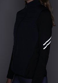 Veste matelassée bleu marine avec un bas noir à manches longues. Présente des accents réfléchissants sur la manche et une fermeture éclair à l'avant. Matière légère.