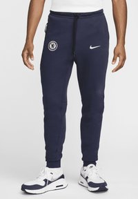 Tummansiniset Nike-housut, joissa on Chelsea FC -logo, kiristysnuora vyötäröllä, vetoketjulliset taskut ja ribattu helmustus, yhdistettynä valkoisiin ja tummansinihin lenkkareihin.