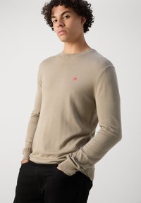 Beiger Strickpullover mit Rundhalsausschnitt, langen Ärmeln und gerippten Bündchen. Mit einem kleinen roten Logo auf der Brust, glatte Textur.