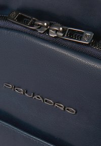 Detalle de bolso de cuero azul marino con cremallera de metal, costuras en contraste y logo en relieve. Textura suave con elementos de diseño elegantes.