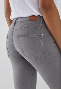 Jeans de mezclilla gris con un ajuste ceñido. Presenta dos bolsillos traseros, un pequeño adorno en el bolsillo y una etiqueta en la cintura.