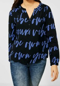 Czarna bluzka z niebieskim wzorem przypominającym ręcznie pisany tekst. Posiada częściowe zapięcie na guziki, długie rękawy oraz luźny krój z delikatnym marszczeniem na ramionach.