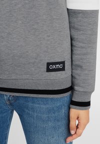 Grå sweatshirt med svarta och vita detaljer, ribbade manschetter och en logotyp-patch. Tyget verkar mjukt, ledig passform. Bärs med blå slitna jeans.