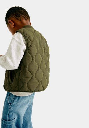 Niño pequeño vistiendo un chaleco acolchado verde oliva sobre una sudadera gris claro y jeans azules, de espaldas sobre un fondo liso.