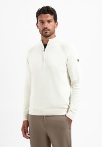Crème-kleurige pullover met een halve rits en ribbelaccenten op de schouders, en een getailleerde zoom. Combineer met beige broek voor een samenhangende look.