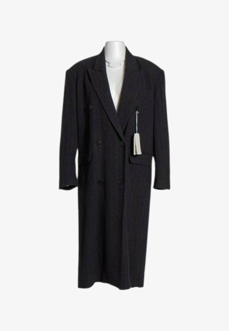 Long manteau noir croisé avec de larges revers, des poches à rabat et une étiquette de prix suspendue sur un buste de mannequin blanc.