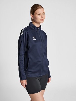 Vrouw draagt een marineblauw sportjack met rits en hummel-logo en zwarte fietsbroekjes, staand tegen een effen lichte achtergrond.