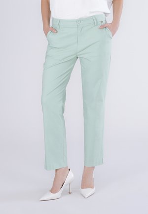 Pantalon vert clair à jambes droites avec poches avant, porté avec des chaussures blanches à talons hauts et un haut blanc rentré.