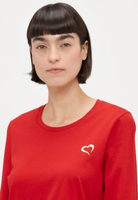 Rotes Langarmshirt aus weicher Baumwolle mit einem goldenen Herz-Graphic auf der linken Brustseite und einem runden Ausschnitt.