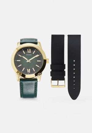Montre-bracelet dorée avec un bracelet en cuir vert foncé, un cadran noir texturé, des chiffres romains, et deux bracelets en caoutchouc noir supplémentaires.