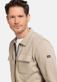 Beige zip-up jack met twee borstzakken en een zwart logo-patch op de schouder. Gemaakt van textuurstof en voorzien van een klassieke kraag.