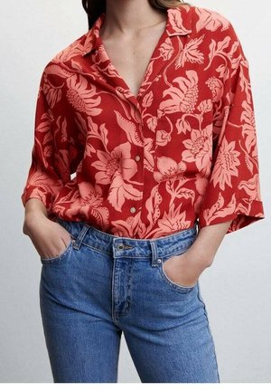 Person iført en rød blomsterprint knappet skjorte med op-rullede ærmer, stoppet ned i højtaljede blå jeans, hænderne i lommerne.