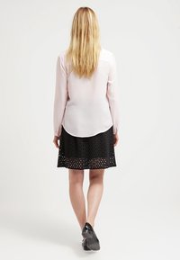 Helle rosa Bluse mit langen Ärmeln und abgerundetem Saum, kombiniert mit einem schwarzen Rock mit geometrischem Ausschnitt. Schwarze Schuhe ergänzen das Outfit.