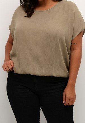 Vrouw in een losse, kort-ge mouwen beige top, in een hoge taille zwarte broek, met een zichtbare tatoeage op haar linkerarmp.
