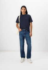 Mørkeblå t-skjorte med korte ermer og en subtil logo, kombinert med lyseblå straight-leg jeans og hvite joggesko, alt på en enkel bakgrunn.