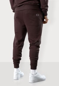 Brown sweatpants made of soft fabric, featuring a fitted design, ribbed cuffs, and a small logo detail on the back pocket. Worn with white shoes.