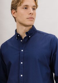 Marineblå skjorte med knapper foran, vanlig krage, hvite knapper og glatt tekstur. Nærbilde av overkroppen og kragen.