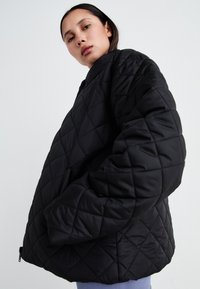 Veste oversize matelassée noire avec une coupe carrée, présentant des motifs en forme de diamant, un col montant et des poches latérales. Texture de tissu lisse et brillant.