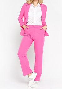 Bright pink suit with tailored jacket and matching trousers, white shirt with dot pattern, and white sneakers with silver accents.