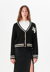 Mustakotoinen neulecardigan, jossa on v-pääntie ja valkoiset ribbireunukset. Varustettu suurilla napeilla ja "NY"-embleemillä. Lyhyempi muotoilu ja pitkät hihat.