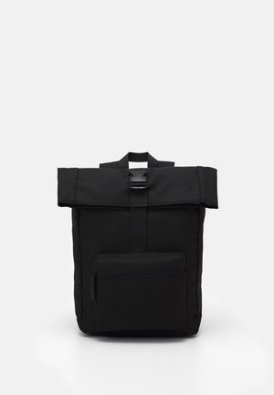 Sac à dos noir à fermeture roulante avec poche avant zippée et fermeture à boucle centrale, debout sur un fond clair uni.